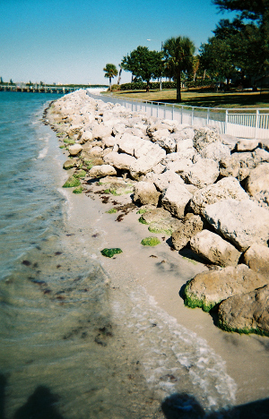 [Standing on a bridge looking to the right and seeing the rocks of the quay and shoreline with a railing and walkway above the quay.]